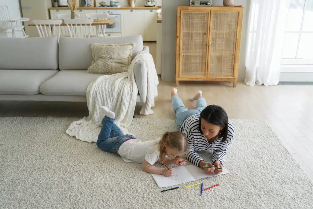 Mamma och dotter ligger på en ljus matta i ett vardagsrum och färglägger tillsammans med färgpennor.
